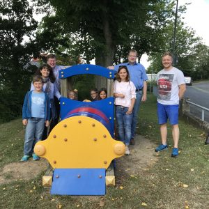 Mehrere Kinder in einer Spiel-Lokomototive, daneben weitere lächelnde Kinder und Erwachsene.